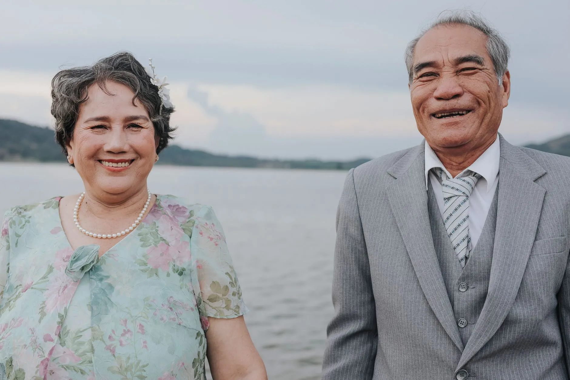 A smiling older Asian couple dressed for a celebration beside the water.