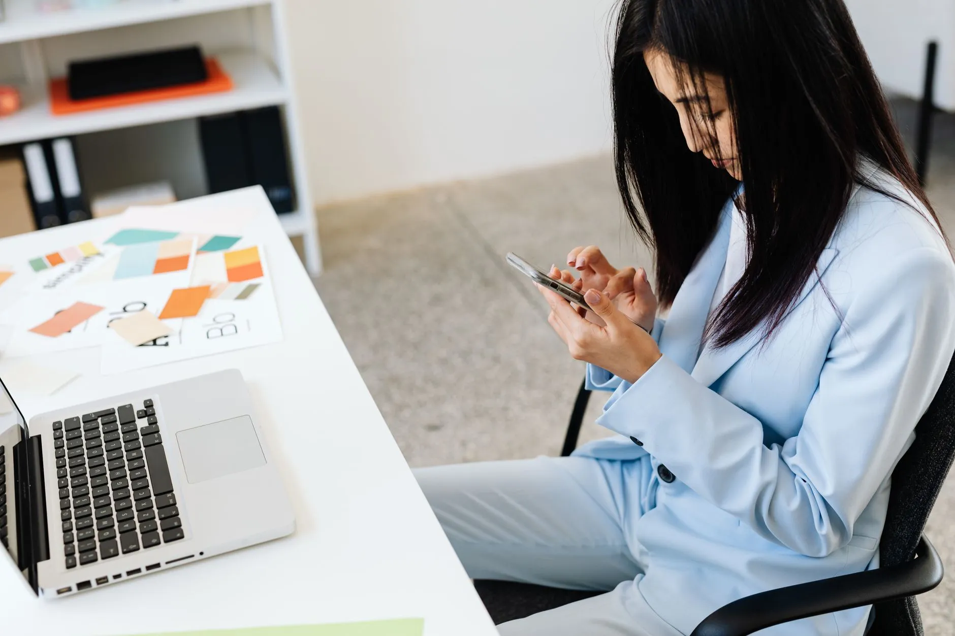 An Asian professional in a blue suit using her smartphone at an office desk.