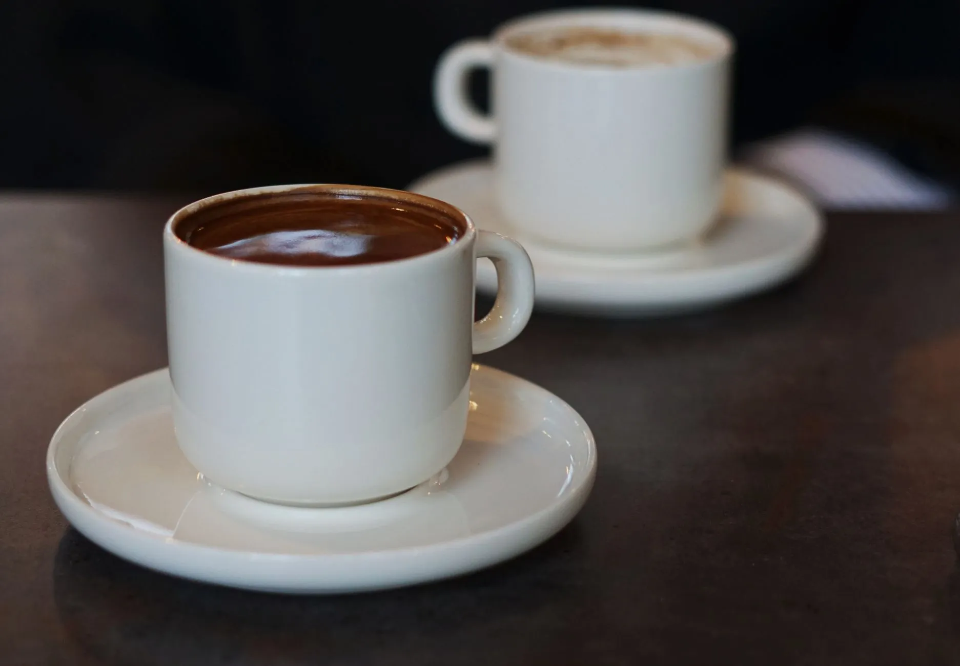 Two white cups of coffee side by side on a café table.