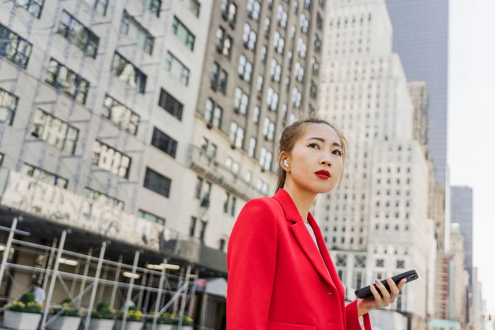 An Asian woman in a red blazer holding a smartphone on a city street.
