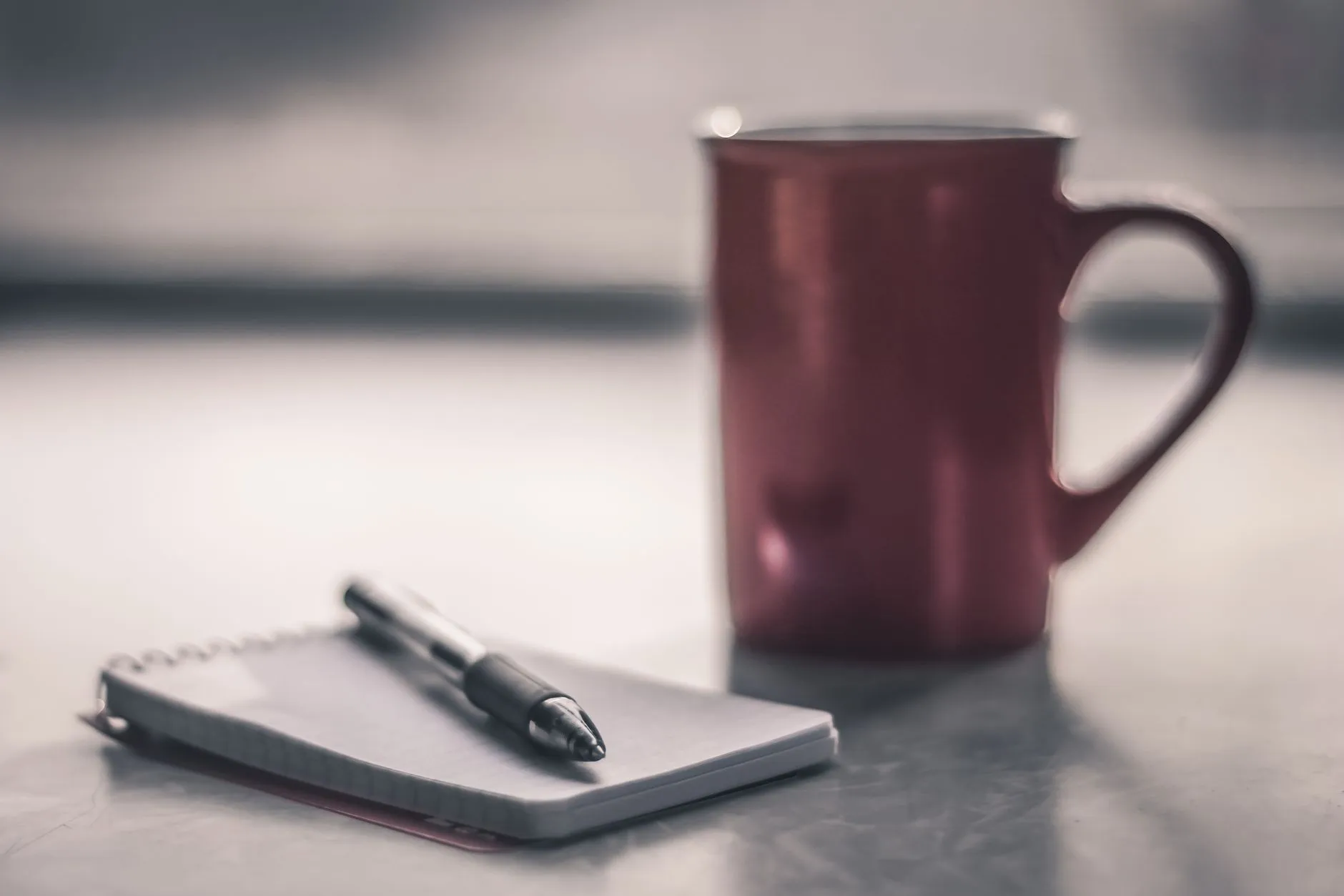 A notebook and pen beside a cup on a desk in soft morning light.