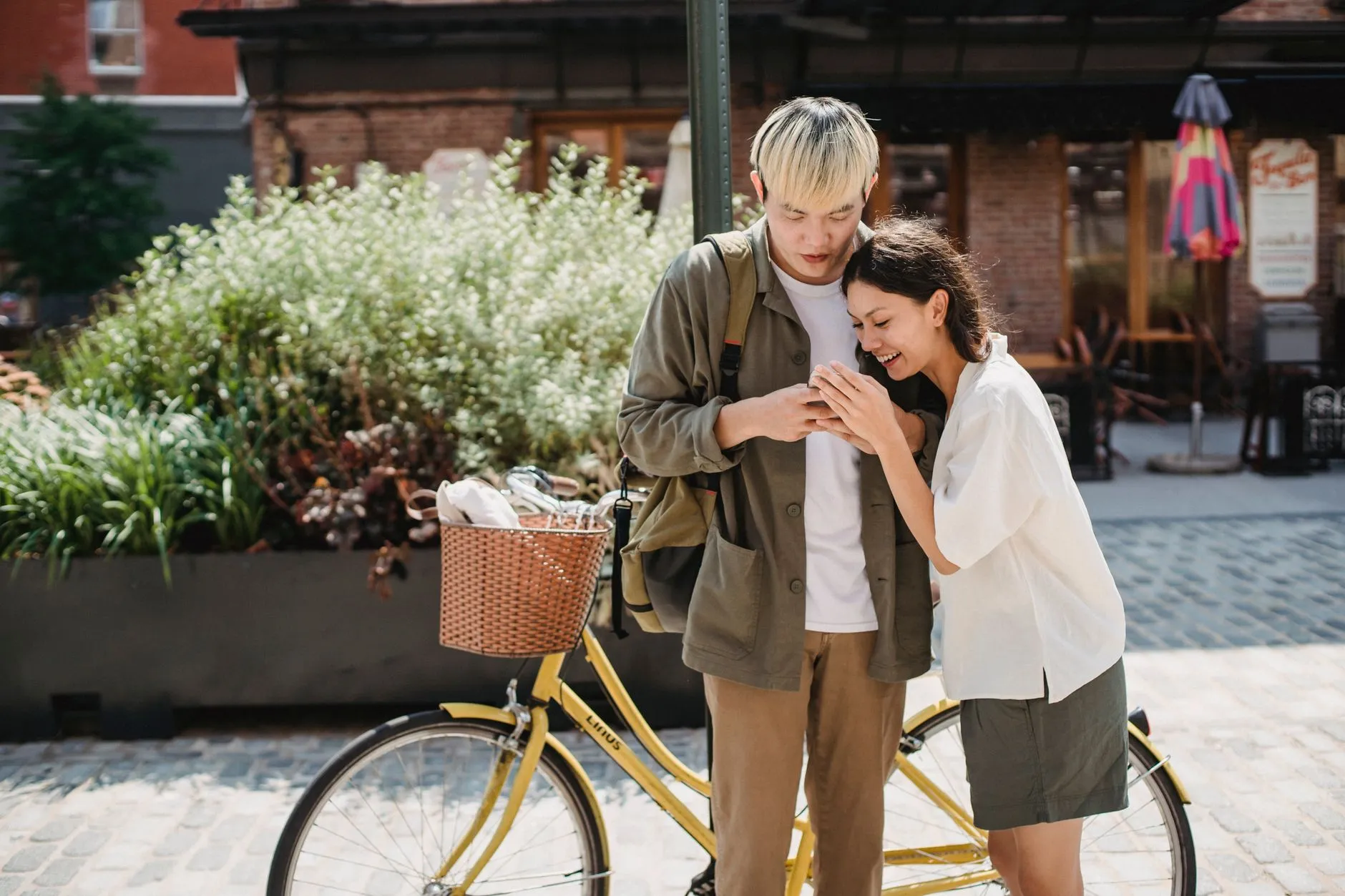 A young Asian couple smiling as they look at a smartphone beside a bicycle.