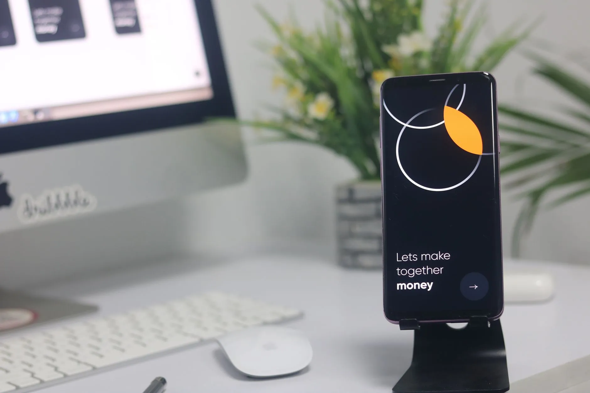 A smartphone on a tidy desk beside a small plant and a computer setup.