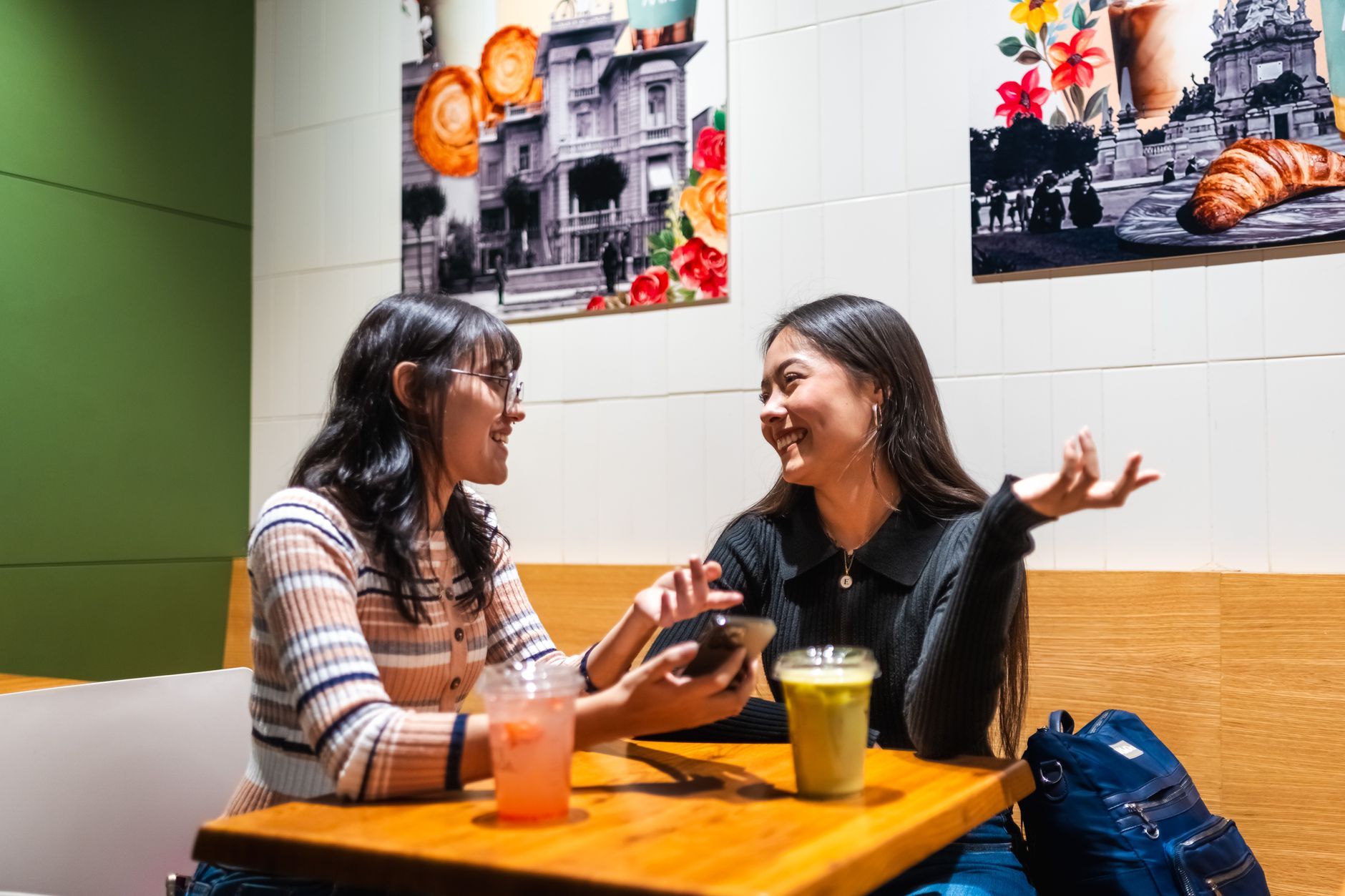 Two Asian people smiling while exchanging messages on their phones at a cafe.