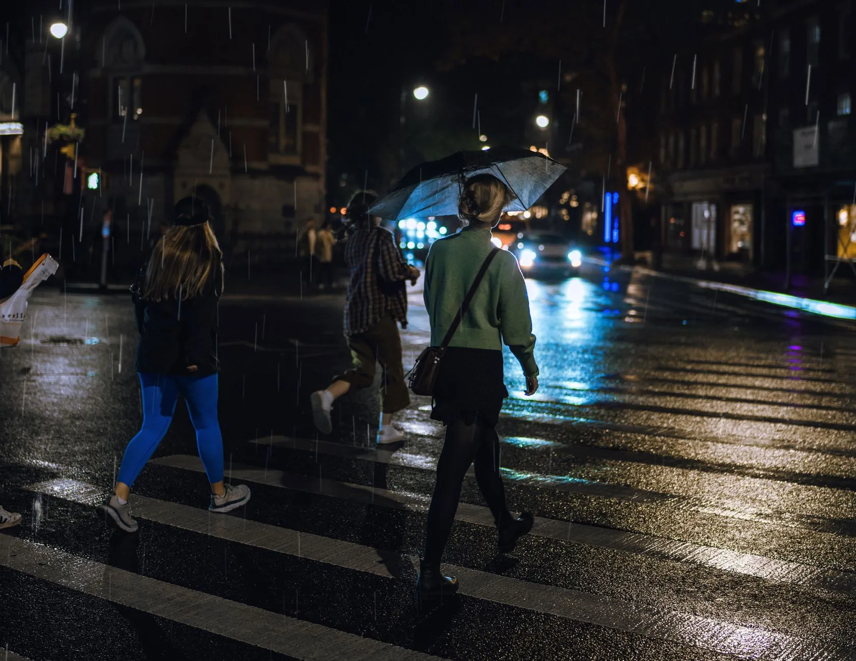 People crossing a wet city street at night under umbrellas, lit by street lights.